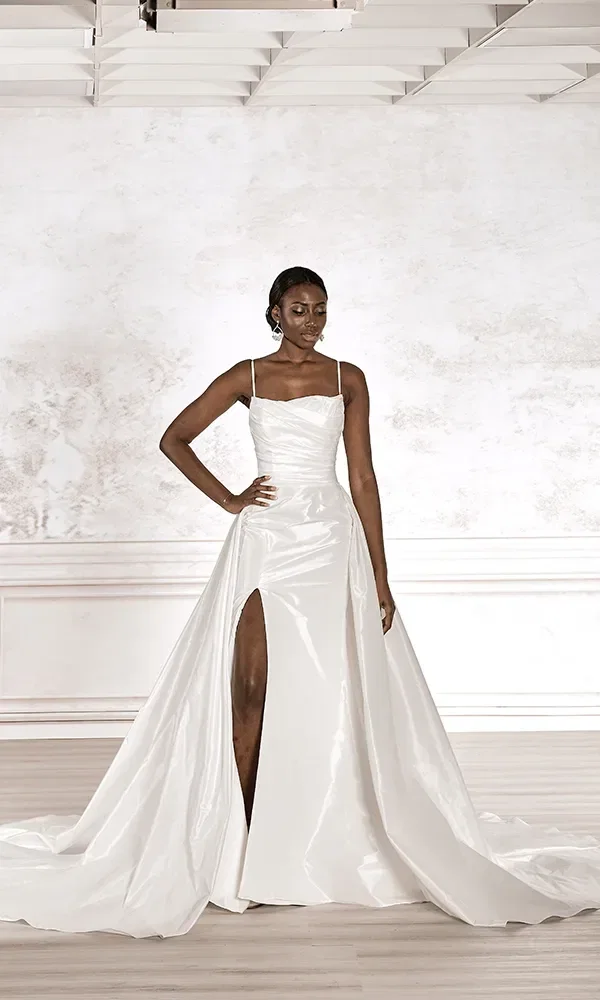 Eine Frau in einem weißen Hochzeitskleid mit hoher Taille und Beinschlitz posiert stehend, ihre Hand ruht lässig auf der Hüfte, vor einem hellen, strukturierten Hintergrund.
