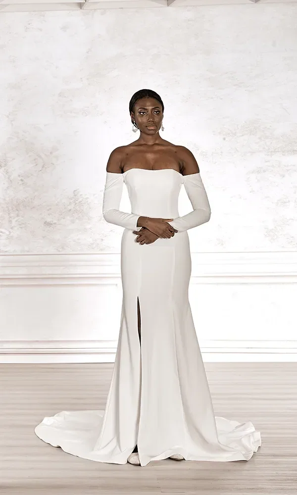 Eine Frau in einem weißen Brautkleid mit langen Ärmeln und schulterfreiem Design steht mit übereinander geschlagenen Händen vor einem hellen Hintergrund.