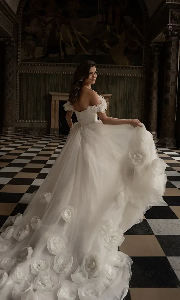 Eine Frau in einem prächtigen weißen Brautkleid mit Rosenapplikationen dreht sich um und blickt über ihre Schulter, stehend vor einem schachbrettartigen Boden und historischen Wandgemälden.
