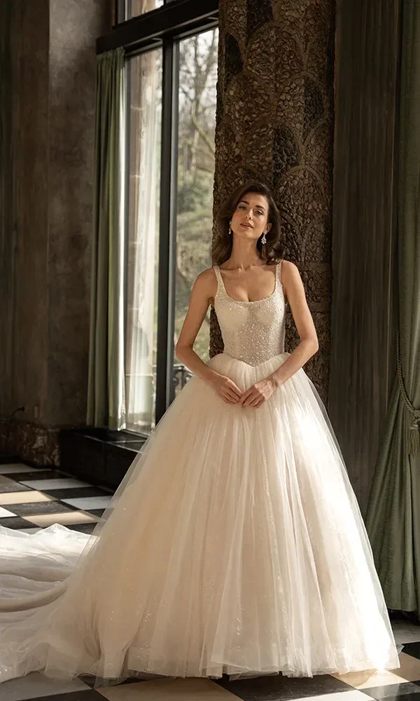 Eine Braut in einem funkelnden ärmellosen Brautkleid mit einem weiten Tüllrock posiert in einem Raum mit großen Fenstern und reich verzierten Vorhängen.