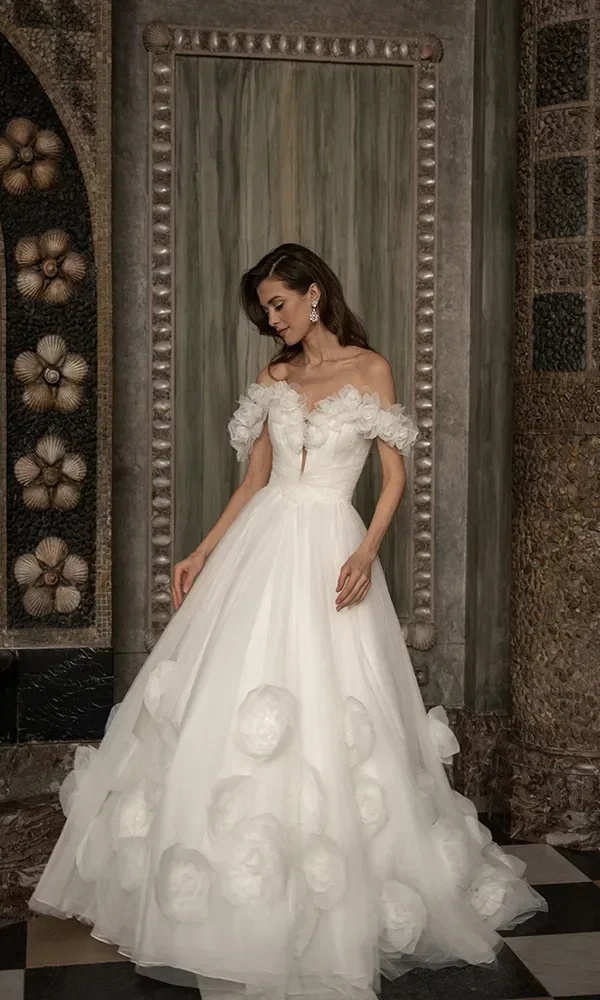 Eine Frau in einem weißen Brautkleid mit Blumenapplikationen und schulterfreiem Design steht vor einer historischen Wand mit kunstvollen Gemälden und Dekorationen.