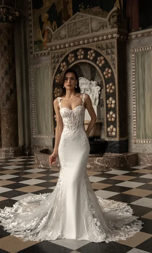 Eine Frau in einem weißen, figurbetonten Brautkleid mit transparenten Details und Blumenapplikationen steht in einem historischen Saal mit schachbrettartigem Boden und verzierten Säulen.