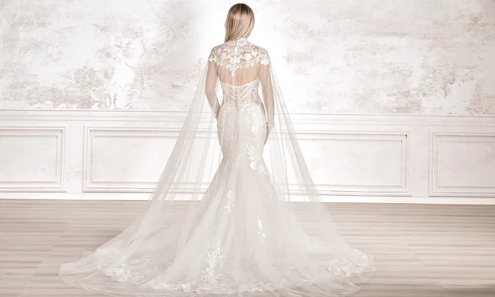 Eine Frau von hinten gesehen im weißen Hochzeitskleid mit aufwendiger Spitze und einer langen, transparenten Schleppe, die sich über den Holzboden eines schlichten Raums erstreckt.