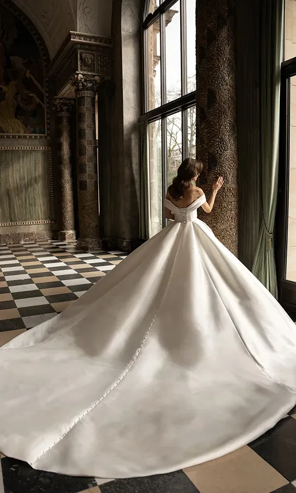 Rückansicht einer Braut in einem weißen Kleid mit langem, mit Spitze besetzten Schleier, die in einem eleganten Raum vor einem Kamin steht.