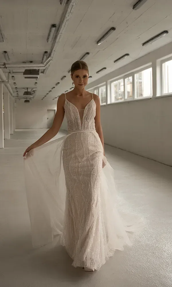 Eine Braut in einem langärmligen Brautkleid mit transparenten Spitzenärmeln und einem weißen Rock steht vor einem hellen Fenster.