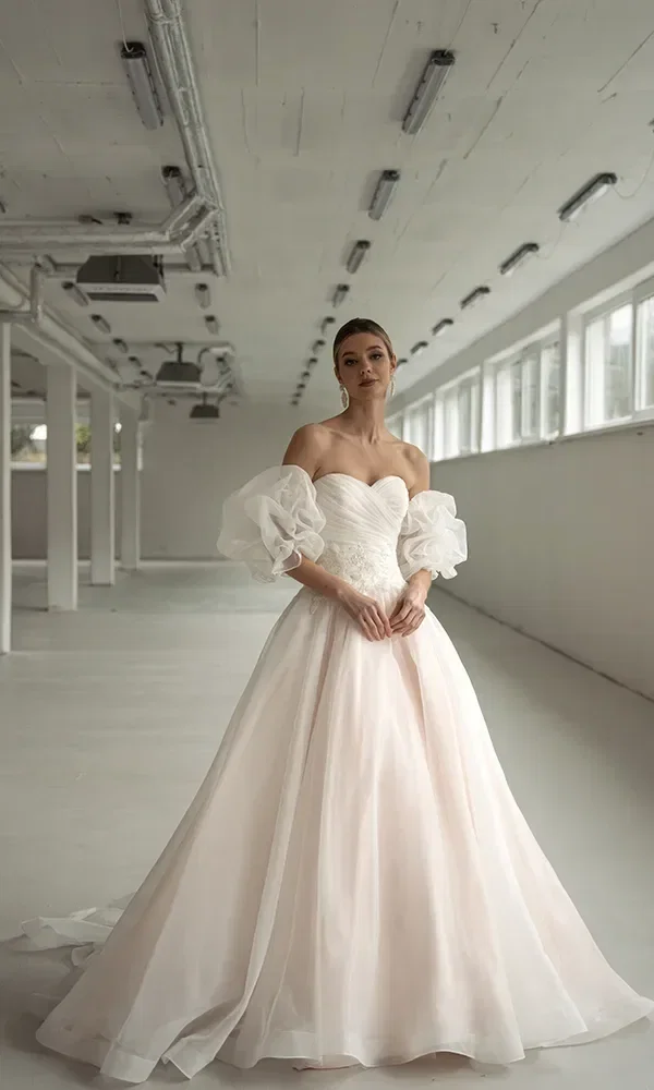 Eine Braut in einem weißen Kleid mit schulterfreien Puffärmeln und einem weiten Rock posiert in einem industriellen Raum.