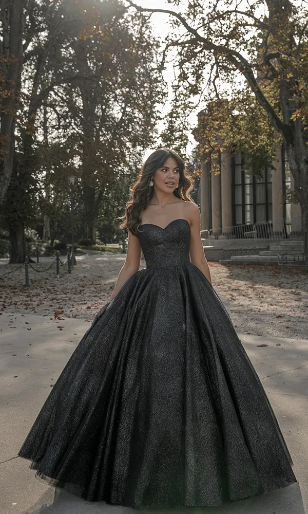 Eine Braut in einem voluminösen schwarzen Ballkleid mit Herzausschnitt steht auf einem Laubweg in einem Park.
