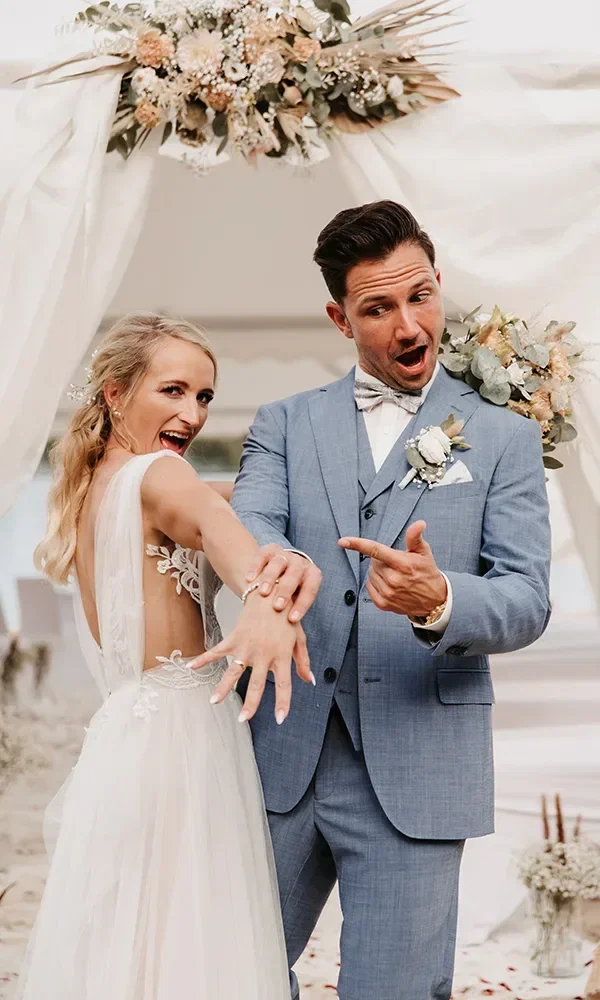 Glückliches Hochzeitspaar zeigt stolz ihre Eheringe, wobei der Bräutigam begeistert auf die Ringe deutet und beide in die Kamera schauen, unter einem blumengeschmückten Hochzeitsbogen am Strand.