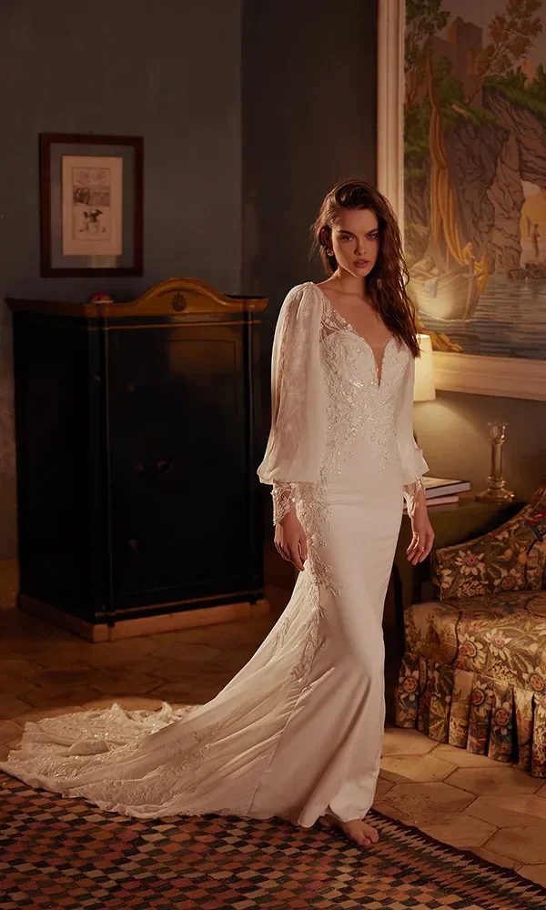 Eine nachdenklich blickende Braut in einem Vintage-inspirierten Kleid mit Rüschensaum und langer Schleppe steht barfuß auf einem orientalischen Teppich in einem historischen Raum.