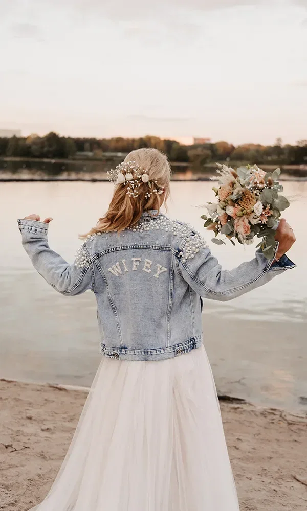 Braut am Strand bei Sonnenuntergang von hinten, mit Blumen im Haar und einer Jeansjacke mit der Aufschrift 'WIFEY' aus Perlen, hält einen Brautstrauß.