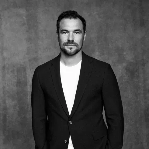 Ein Porträt in Schwarz-Weiß eines Mannes mit kurzem dunklen Haar und leichtem Bart, der ein gut sitzendes dunkles Sakko über einem weißen T-Shirt trägt. Er blickt ernst in die Kamera.