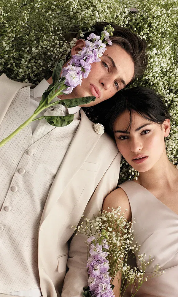 Ein Bräutigam und eine Braut liegen auf einer Wiese mit weißen Blüten, der Bräutigam hält einen lila Blumenstängel vor sein Gesicht. Er trägt einen elfenbeinfarbenen Anzug, sie ein beiges Kleid.