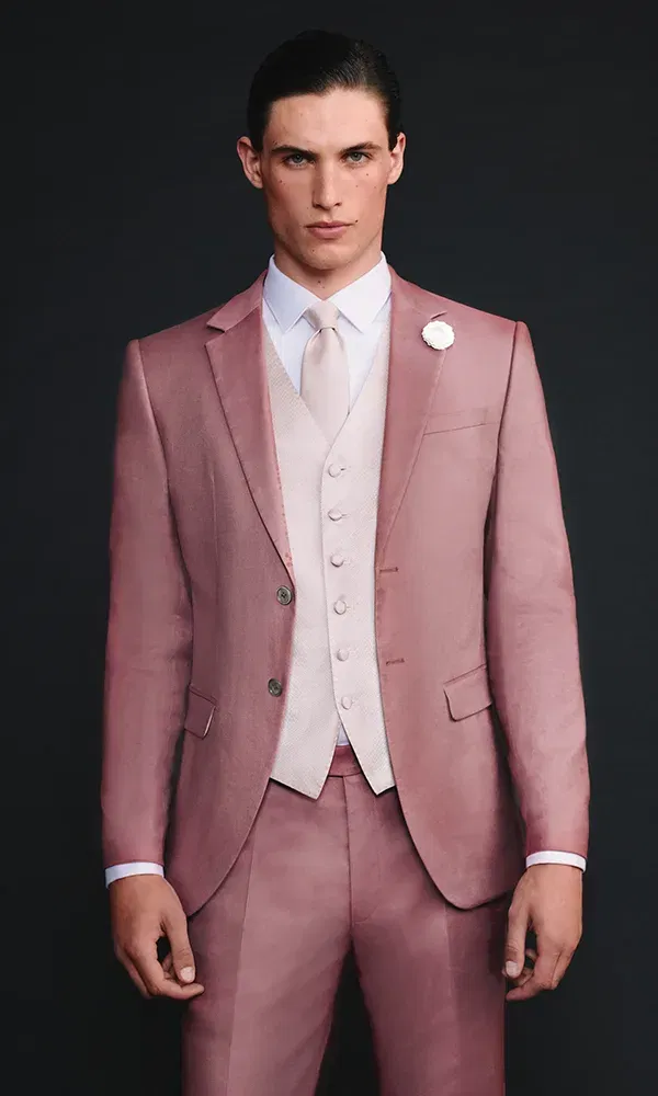 Ein Model in einem rosafarbenen Anzug mit weißem Hemd, passender Weste und Krawatte. Ein weißer Blumenanstecker ziert das Revers. Er steht vor einem dunklen Hintergrund und schaut direkt in die Kamera.