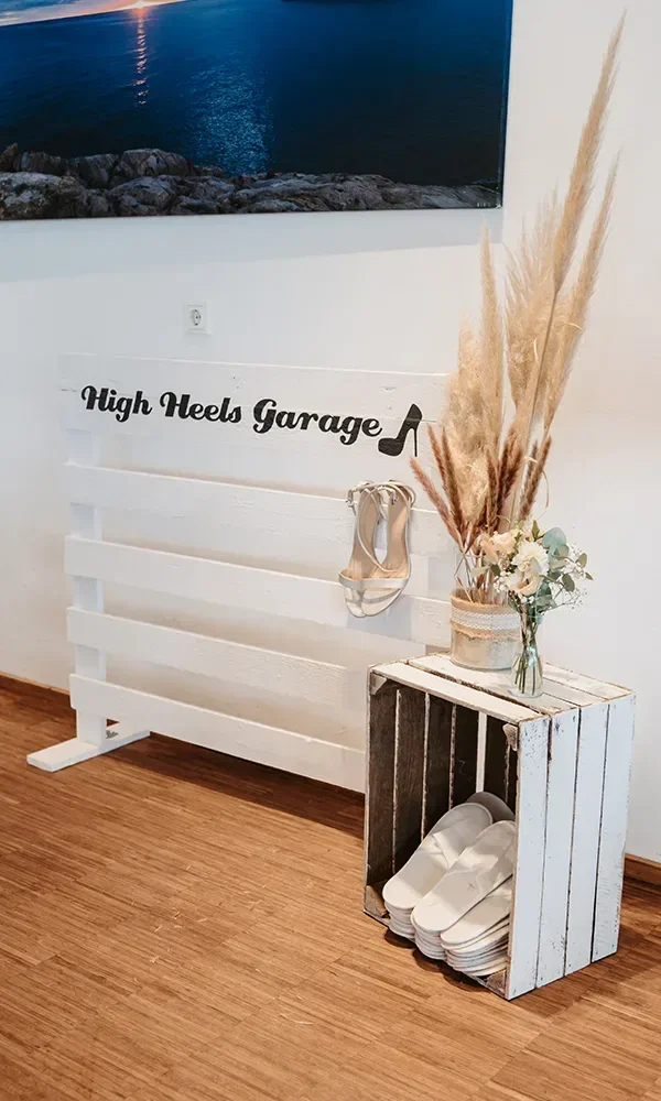 Eine 'High Heels Garage' mit einem rustikalen Holzschild, Brautschuhen auf einer Bank und Flip-Flops in einer Holzkiste für müde Tanzfüße.