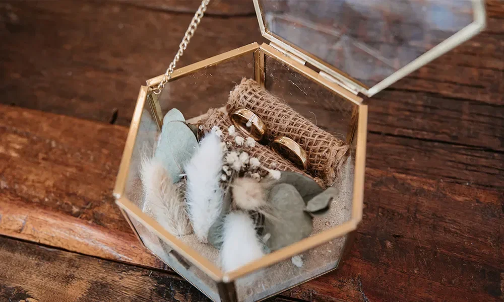 Ein geometrisches Ringkästchen aus Glas mit zwei Eheringen, umgeben von Federn, Eukalyptus und Jute auf einem hölzernen Untergrund.