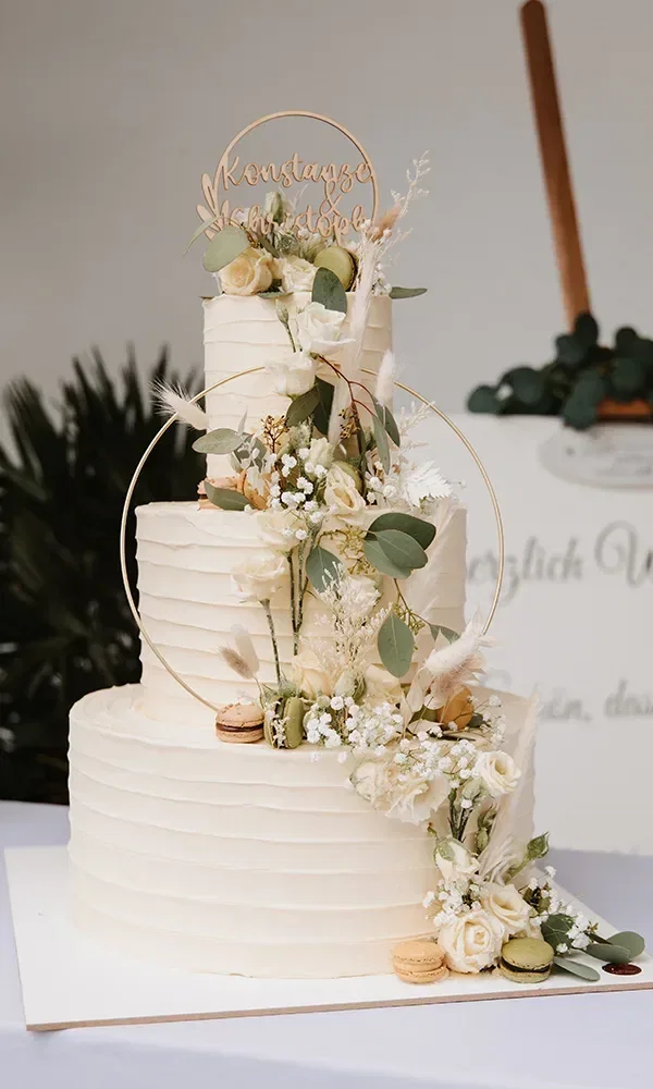 Mehrstöckige Hochzeitstorte, verziert mit frischen Blumen und Macarons, und einem personalisierten Tortentopper mit den Namen 'Konstanze & Christoph' in goldener Schrift.