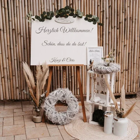 Willkommensschild für eine Hochzeit mit der Aufschrift 'Herzlich Willkommen! Schön, dass du da bist!' neben Namen 'Konzi & Chris' und einem Herz, umgeben von Eukalyptuszweigen.