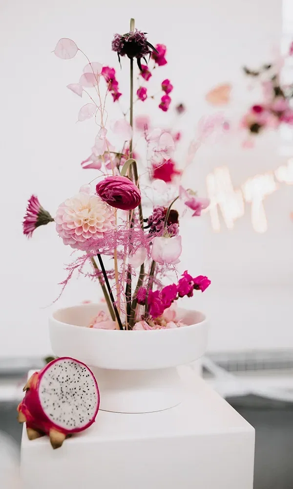 Filigranes Blumengesteck in Pink und Violett in einer weißen Vase, dekoriert mit Drachenfrucht
