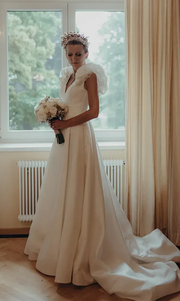 Braut in einem eleganten weißen Hochzeitskleid mit Rüschen und Blumenkrone vor einem Fenster