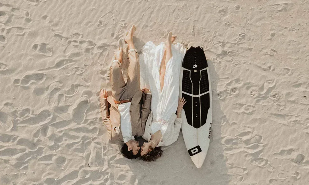 Luftaufnahme eines Hochzeitspaares, das neben einem Surfbrett am Strand liegt