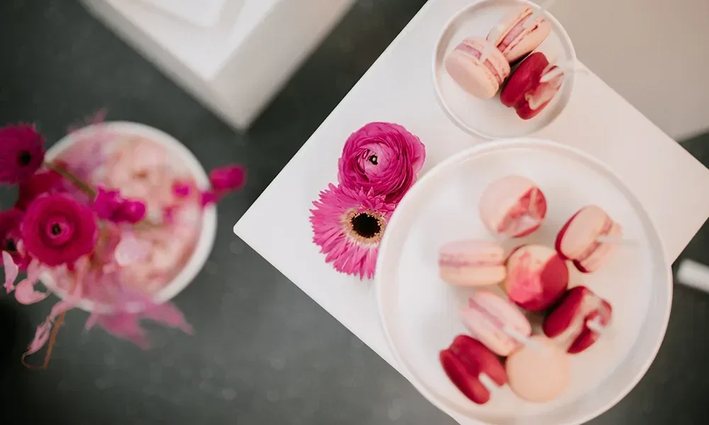 Rosa und pinke Macarons in einer weißen Schale, daneben Blumen in ähnlichen Farbtönen