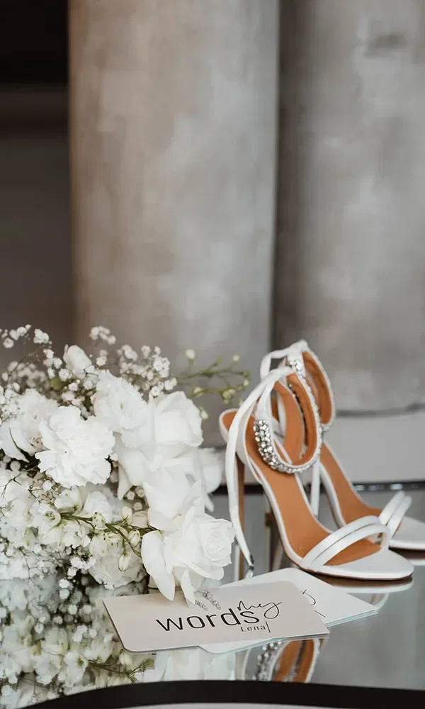 Weiße Brautschuhe mit Strass und ein weißer Blumenstrauß auf einem Tisch