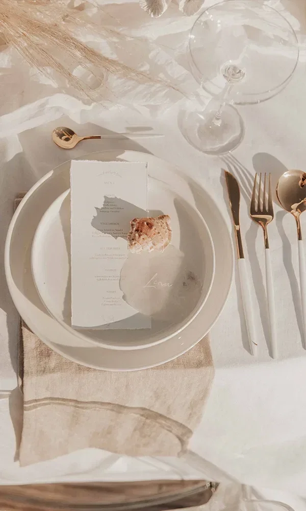 Elegantes Table Setting bei einer Strandhochzeit mit weißen und goldenen Akzenten und einem Menü auf einem weißen Teller