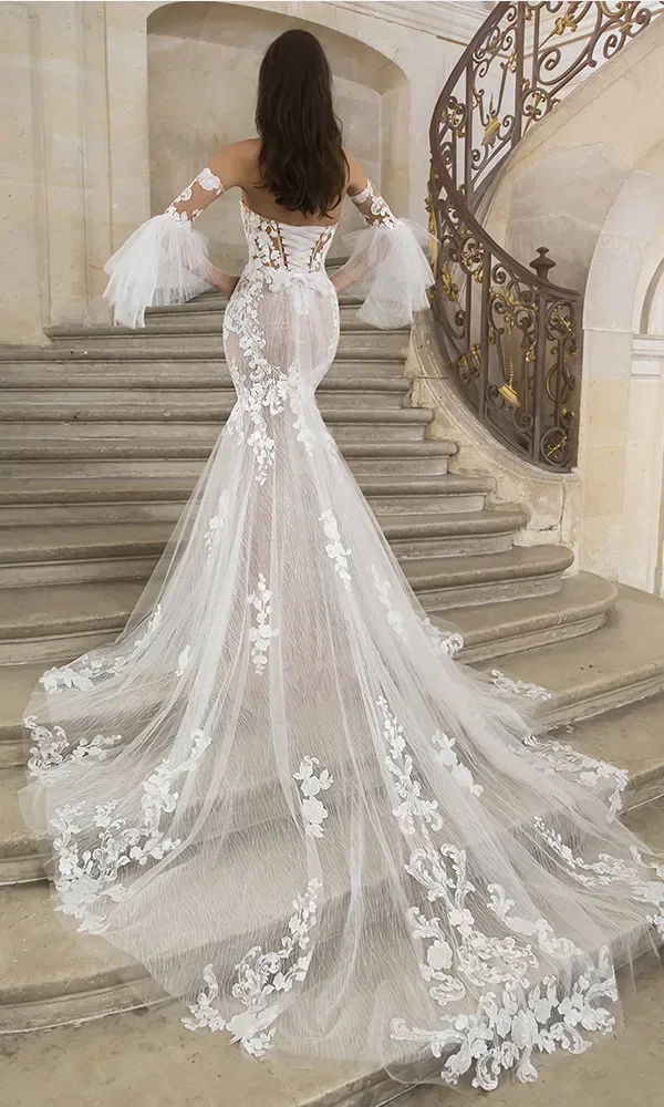Rückansicht einer Braut in einem weißen, bestickten Kleid mit langen transparenten Ärmeln und langer Schleppe, die eine Treppe hinaufgeht.