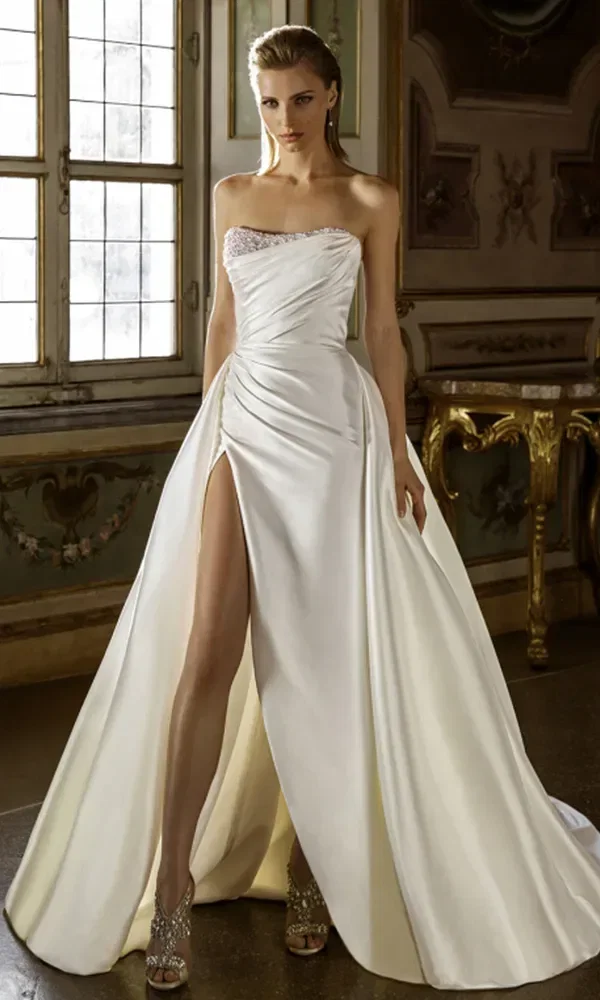 Braut in einem weißen Kleid mit transparentem, besticktem Oberteil und langen Trägern, die in einem reich verzierten Raum steht.