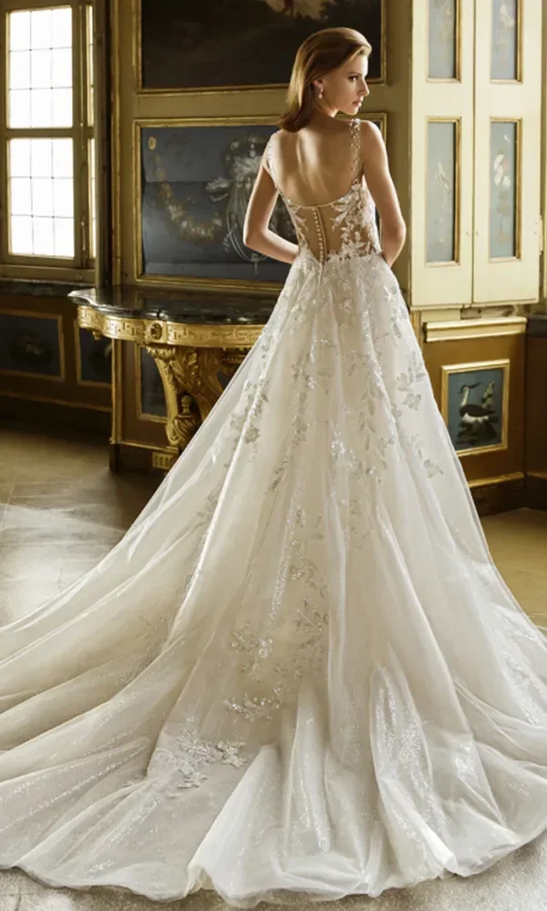 Braut in einem weißen, trägerlosen Kleid mit floralen Applikationen und langer Schleppe, die in einem reich verzierten Raum an einem Tisch lehnt.