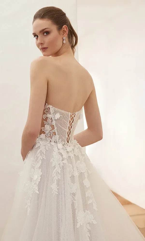 Braut in einem trägerlosen weißen Kleid mit floraler Spitze, die den Rücken zeigt, während sie über die Schulter blickt.