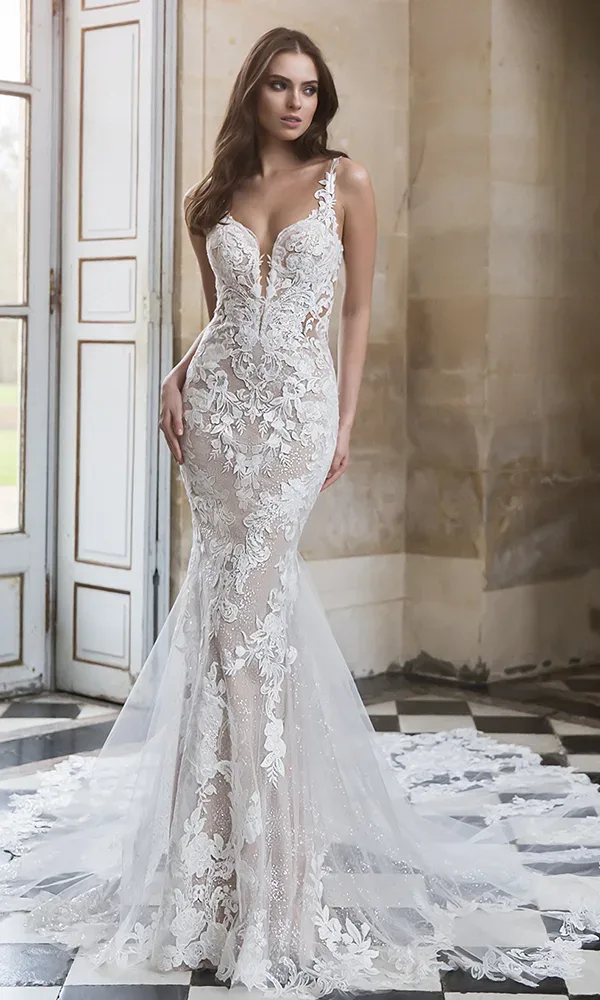 Braut in einem weißen, figurbetonten Kleid mit floralen Applikationen, die in einem eleganten Raum mit großen Fenstern steht.