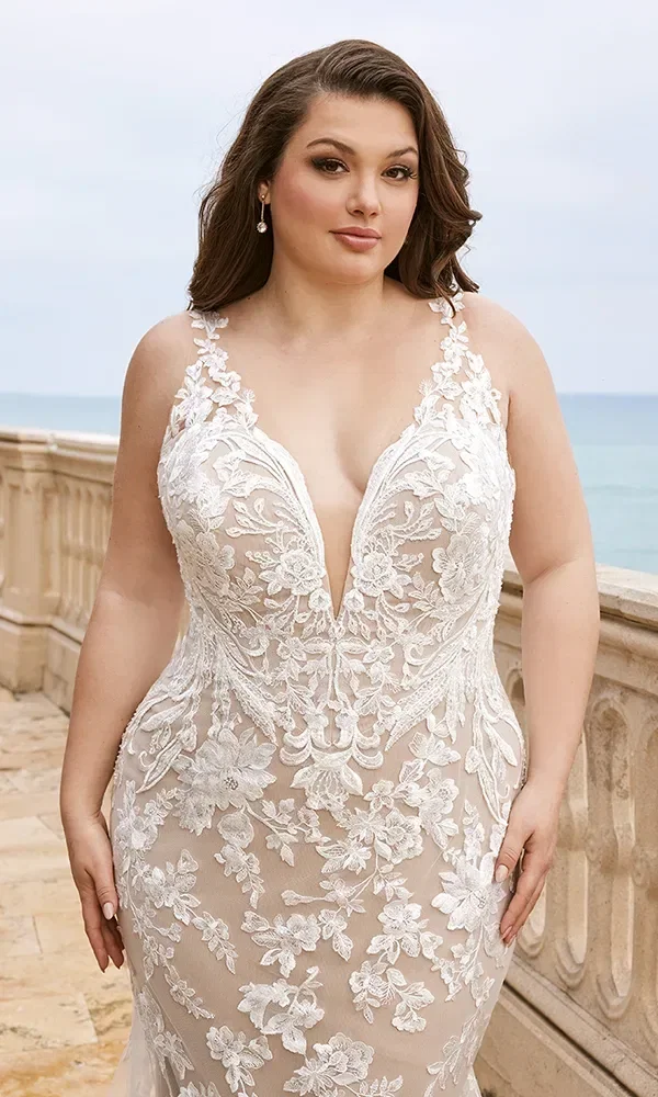 Braut in einem weißen, bestickten Kleid mit tiefem V-Ausschnitt und floralen Applikationen, die draußen an einem Geländer steht.