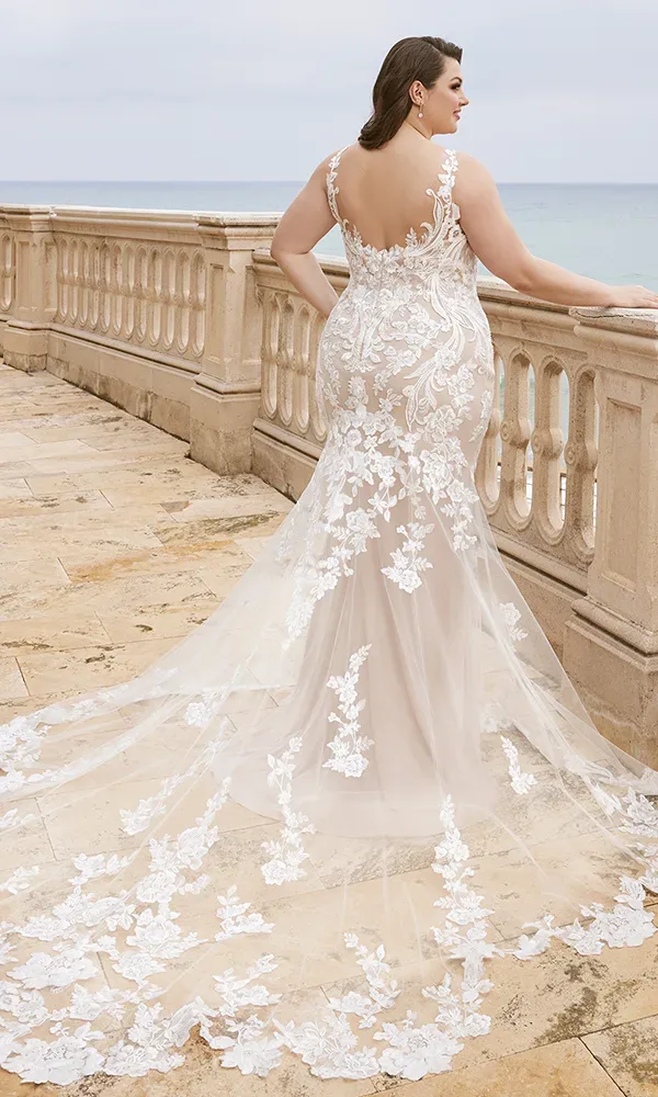 Rückansicht einer Braut in einem weißen, bestickten Kleid mit floralen Applikationen und langer Schleppe, die an einem Geländer am Meer steht.