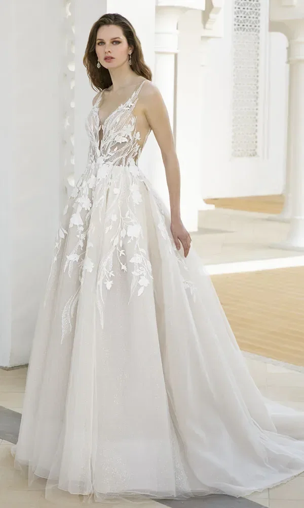 Braut in einem aufwendig bestickten weißen Kleid mit tiefem V-Ausschnitt und dünnen Trägern, die in einem eleganten, weißen Gebäude steht.