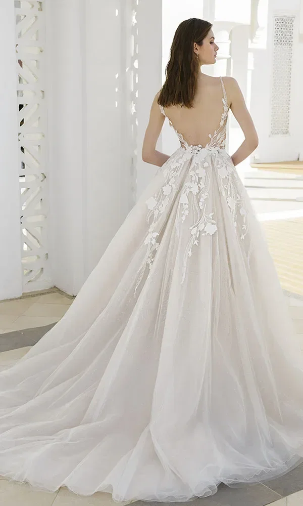 Rückansicht einer Braut in einem weißen, bestickten Kleid mit tiefem Rückenausschnitt und langer Schleppe, die in einem eleganten, weißen Gebäude steht.