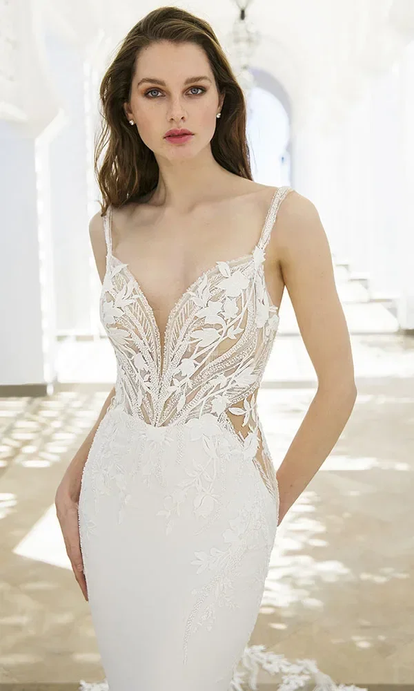 Braut in einem weißen, figurbetonten Kleid mit transparenten Seiten und floralen Applikationen, die in einem eleganten, weißen Gebäude steht.