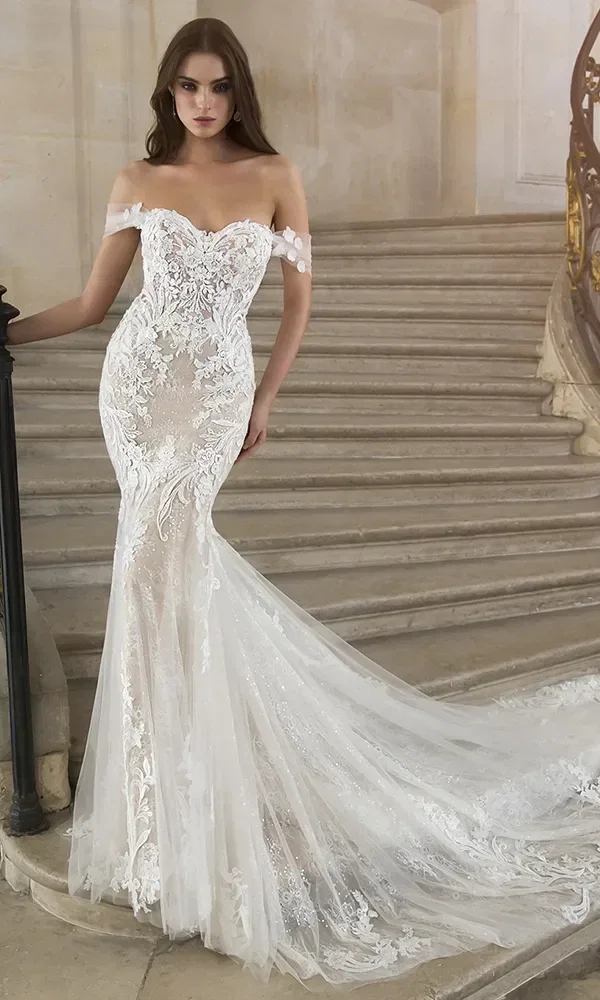 Braut in einem weißen, figurbetonten Kleid mit floralen Applikationen und langer Schleppe, die in einem eleganten Raum steht.