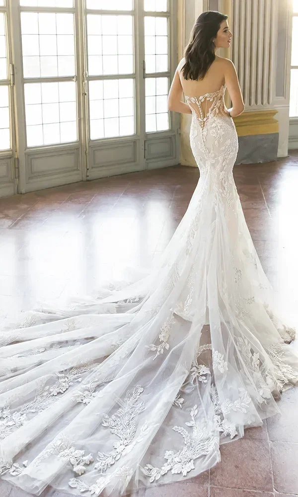 Rückansicht einer Braut in einem weißen, bestickten Kleid mit langer Schleppe, die in einem Raum mit großen Fenstern steht.