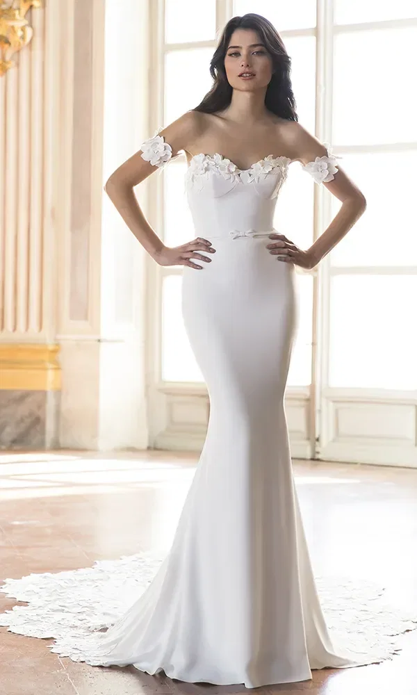 Braut in einem weißen, figurbetonten Kleid mit schulterfreien floralen Applikationen, die in einem eleganten Raum mit großen Fenstern steht.
