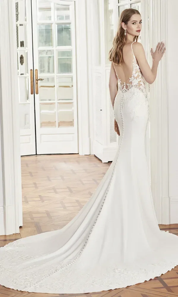 Rückansicht einer Braut in einem figurbetonten weißen Kleid mit tiefem Rückenausschnitt und langen Knöpfen, die in einem eleganten Raum mit Holzboden steht.