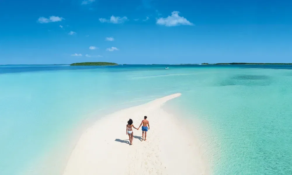 Mann und Frau in Badesachen laufen eine Sandzunge entlang. Drumherum türkises Meer.
