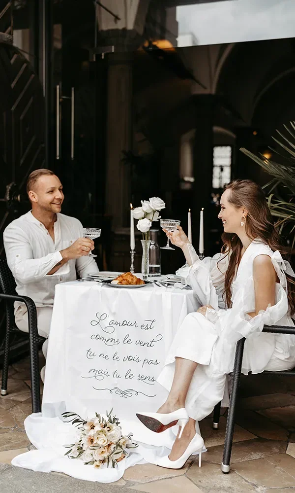 Ein paar sitzt in einem Café an einem gedeckten Tisch mit weißen Blumen, Kaffee und Croissant. Die beiden stoßen mit Champagnergläsern an.