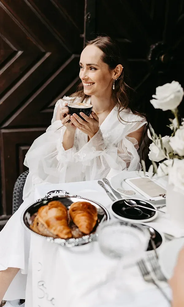 Ein gedeckter Tisch mit weißen Blumen, Kaffee und Croissant, eine Frau in einem weißen Jumpsuit sitzt an dem Tisch und trinkt Kaffee
