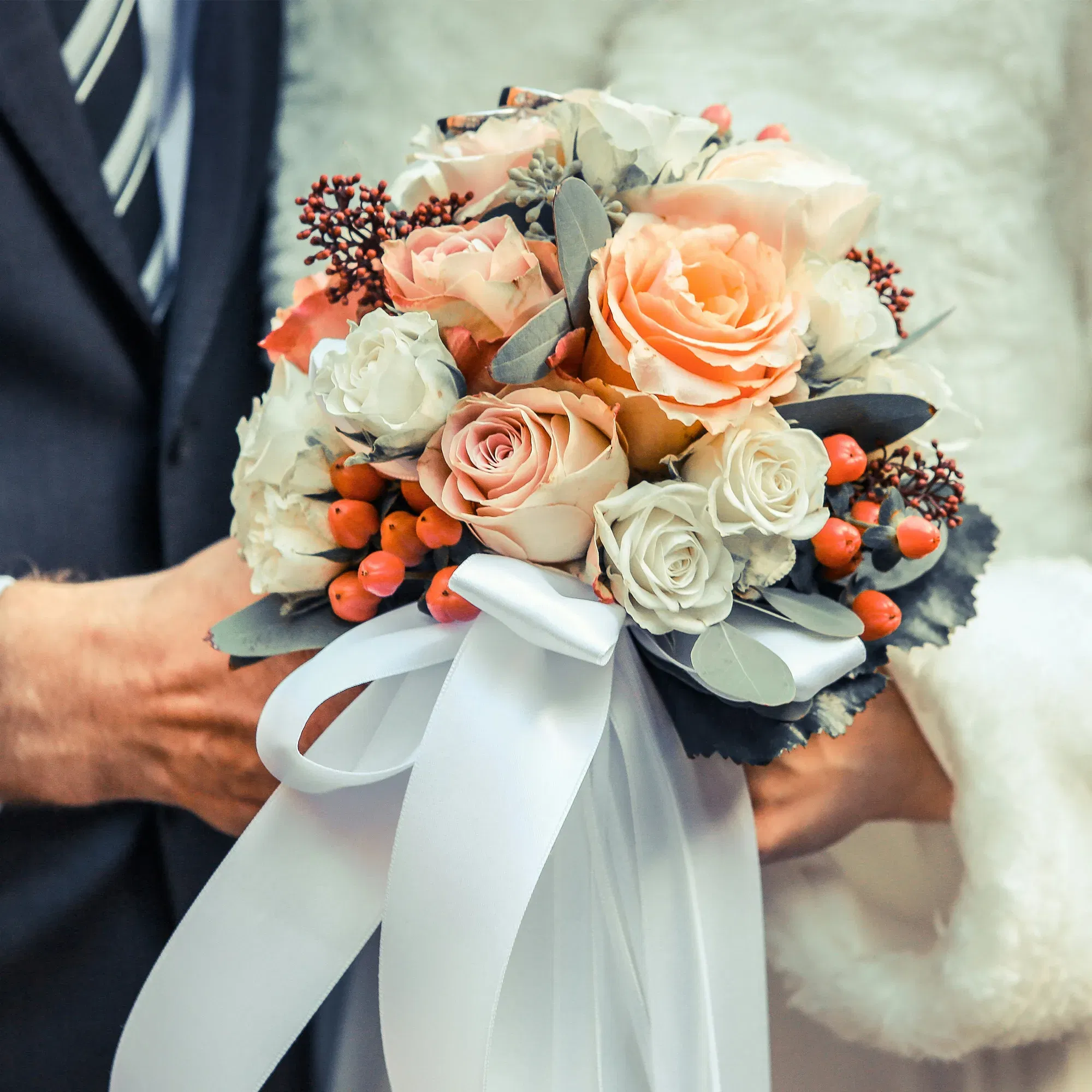 Ein Brautstrauß, gehalten von einem Brautpaar. Der Strauß besteht aus hellrosa und weißen Rosen, orangefarbenen Beeren und graugrünen Blättern. Er ist mit einer weißen Schleife gebunden. Die Hände des Paares sind teilweise sichtbar, die Frau trägt ein weißes, flauschiges Kleidungsstück, und der Mann trägt einen dunklen Anzug mit einer gestreiften Krawatte.