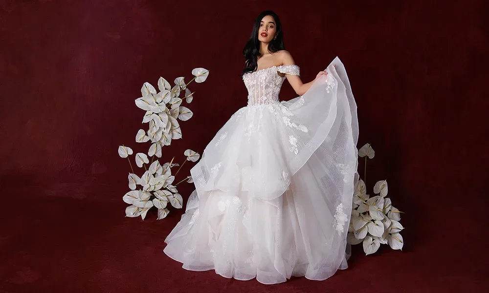 Eine Braut trägt ein schulterfreies Hochzeitskleid mit einem Korsett-Oberteil und einem voluminösen Tüllrock. Sie hält den Rock mit einer Hand hoch, während sie vor einem tiefroten Hintergrund steht, umgeben von weißen Blumen.