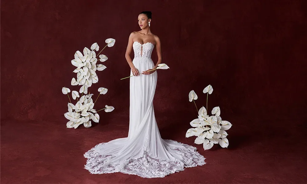 Eine Braut steht in einem schlanken Spitzenhochzeitskleid mit herzförmigem Ausschnitt und langer Schleppe. Sie hält eine einzelne weiße Blume und ist von weißen Blumenarrangements umgeben, vor einem tiefroten Hintergrund.