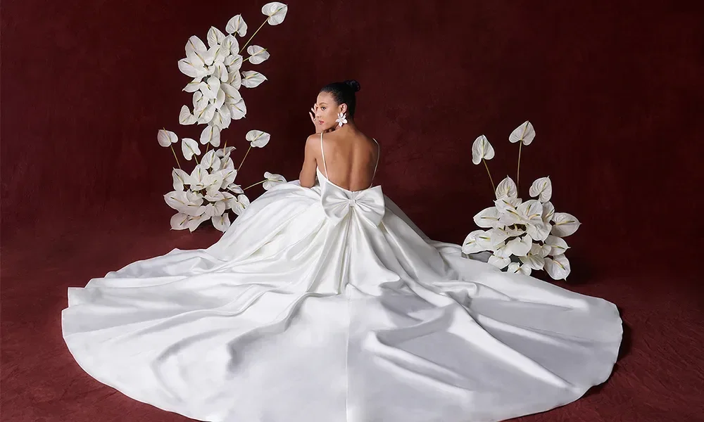Eine Braut sitzt vor einem tiefroten Hintergrund in einem Hochzeitskleid mit einer großen Schleife am Rücken und einer langen Schleppe. Um sie herum sind weiße Blumenarrangements arrangiert.