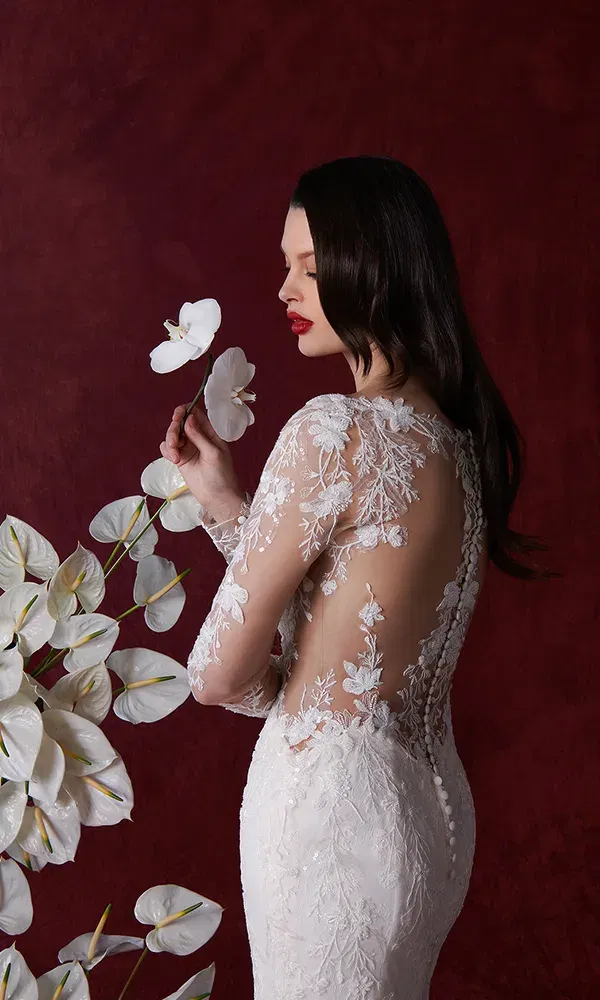 Eine Braut in einem Rückenfreies Spitzenhochzeitskleid, steht vor einem tiefroten Hintergrund und hält eine weiße Blume in der Hand. Im Hintergrund sind weitere weiße Blumen zu sehen.