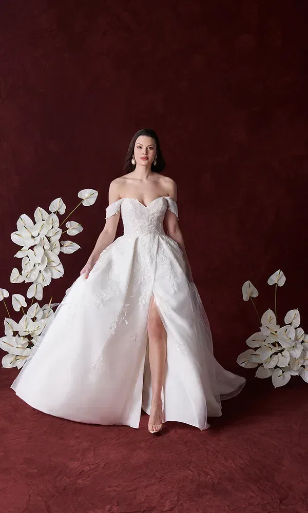 Eine Braut in einem schulterfreien Hochzeitskleid mit einer hohen Schleppe und einem tiefen Ausschnitt. Sie steht vor einem tiefroten Hintergrund, umgeben von weißen Blumen.